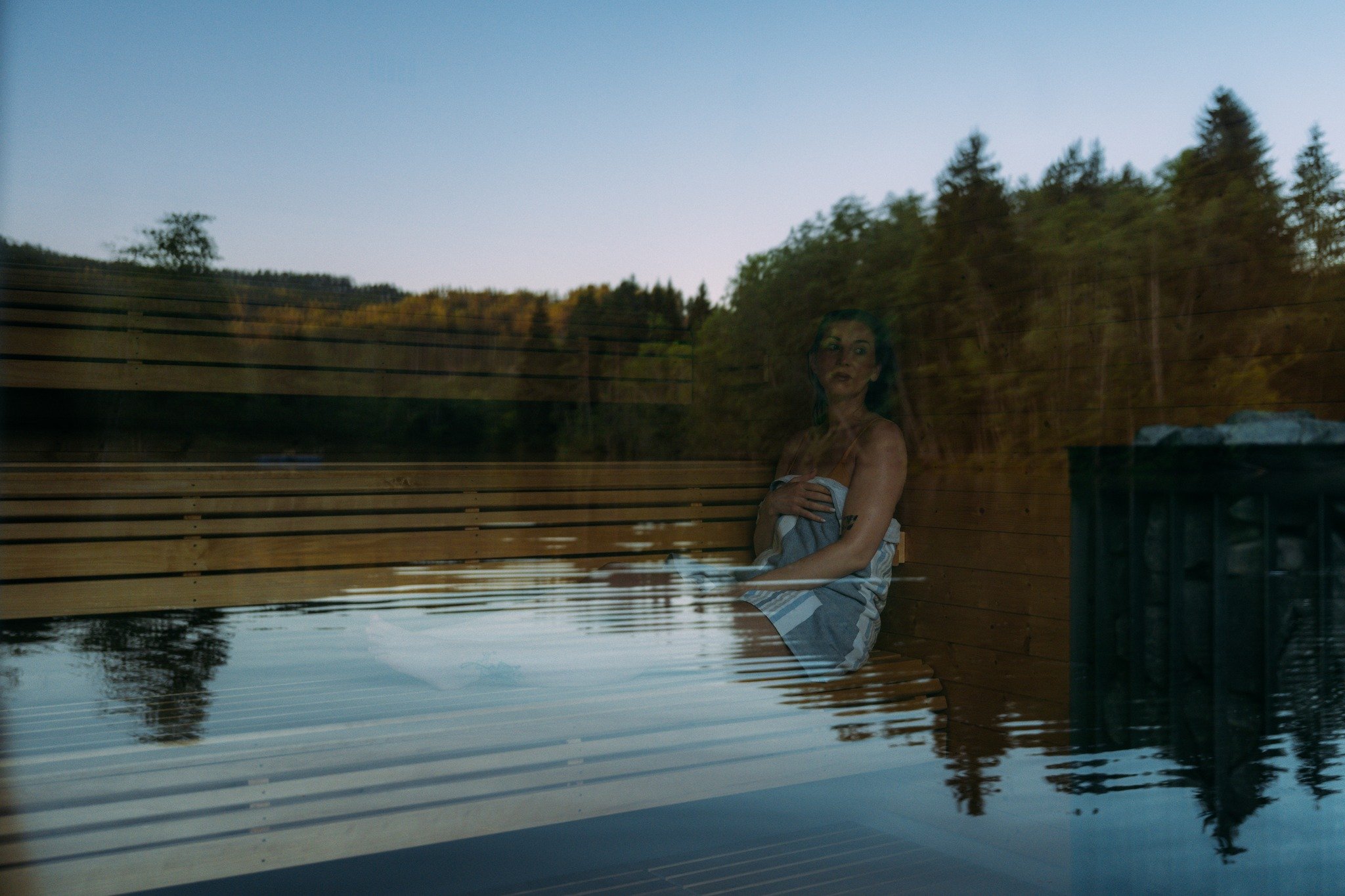 Kvinne sitter i badstue omgitt av naturen, reflektert i glassvindu ved solnedgang.