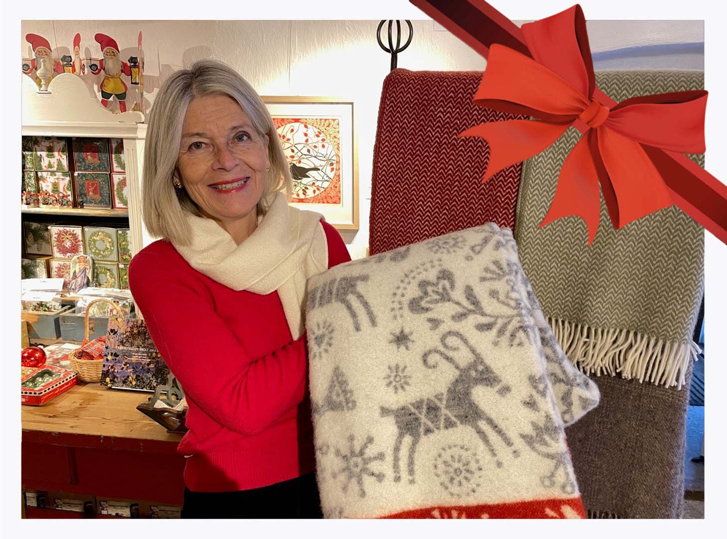 Kvinne i rødt og hvitt viser fram julepledd med reinsdyrmønster i koselig butikkdekorasjon.