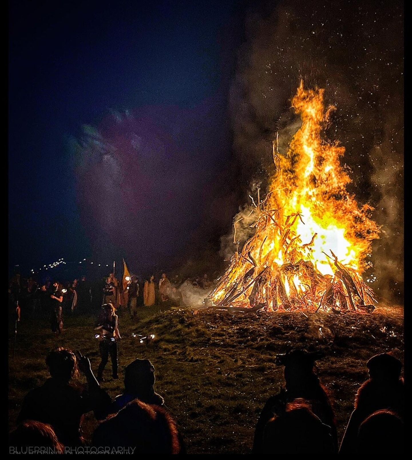 Stor bålbrann på en mørk natt, med mennesker samlet rundt flammene i en festlig atmosfære.