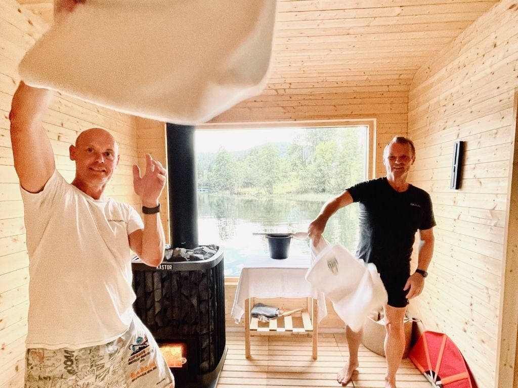 To personer i en badstue kaster håndklær, med utsikt til vann og natur utenfor vinduet.